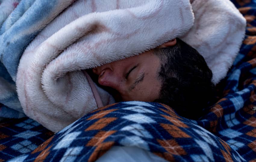 Persona migrante duerme envuelta en mantas en una playa de Ceuta